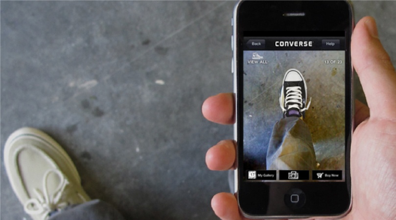 A hand is holding an Iphone and the screen is showing a VR app that is projecting a Converse shoe onto the person’s right feet. The person’s left feet is also visible.