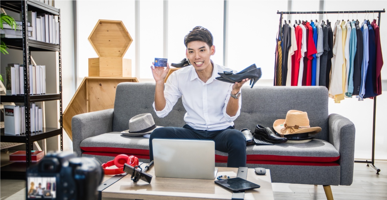 A man sitting on a couch is holding up a credit card in one hand and a pair of ladies’ shoes in the other while sitting in front of a laptop. A DSLR camera is seen videotaping him and various other fashion items are placed around him