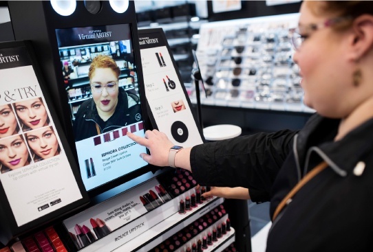 A woman is using a touchscreen display that is placed on a makeup display counter. Her face is virtually mirrored on the screen