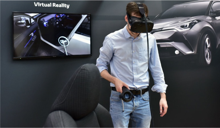 A man is wearing a Virtual Reality headset while holding a VR handheld controller. A TV screen behind him is showing him seeing a VR car with the driver seat door opened