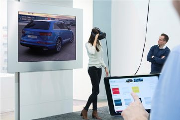 A woman is wearing a VR headset and a screen behind her is showing her seeing a blue SUV. A man is looking at her with his arms crossed. Another person is seen operating an Ipad at the corner of the room.